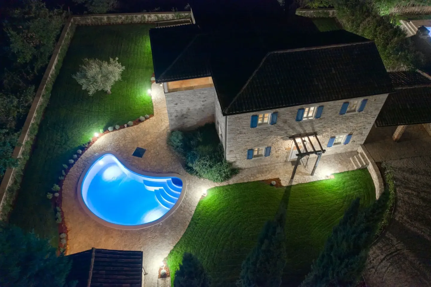 Aerial night view of a house with a lit swimming pool and garden lights.